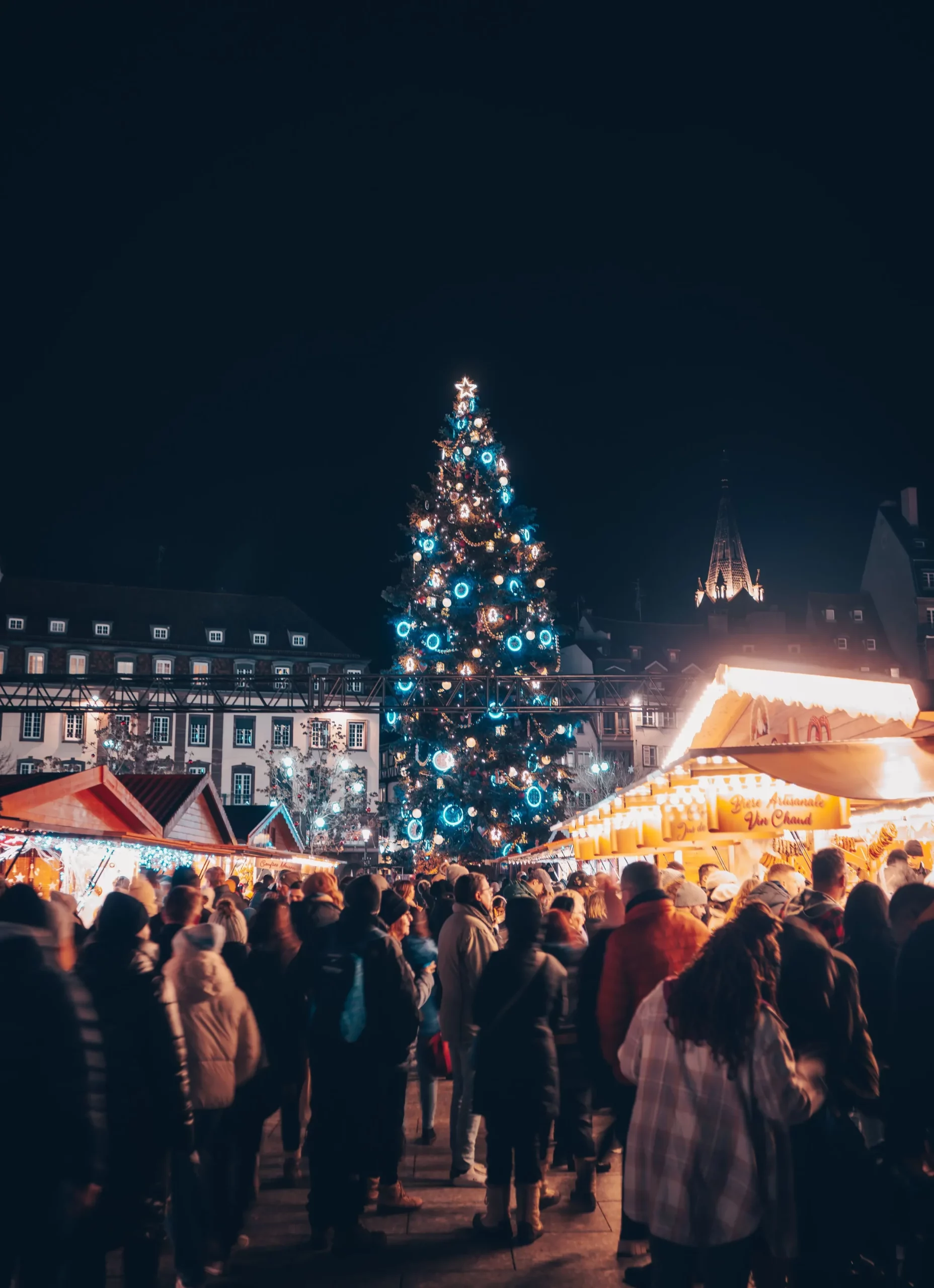 Night-time view of the bustling Strasbourg Christmas market, where a massive, brightly lit Christmas tree rises above warm, glowing wooden stalls and crowds in winter clothing